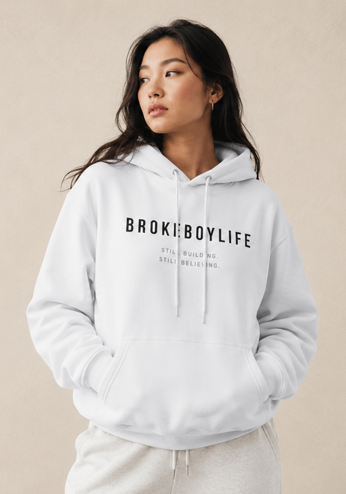 Woman wearing the white BROKEBOYLIFE hoodie featuring the "Still Building. Still Believing." front graphic.