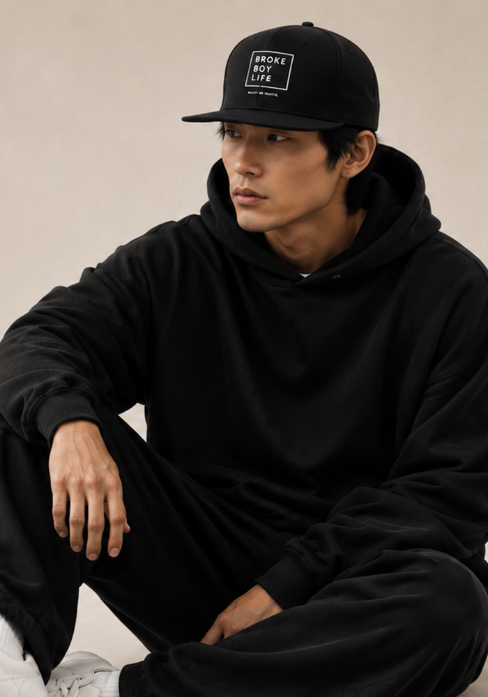 Model wearing a black BROKEBOYLIFE snapback hat with minimalist logo and black hoodie in a lifestyle portrait.