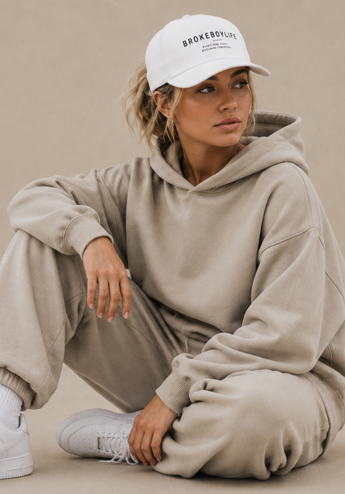Woman wearing a white BROKEBOYLIFE baseball cap with a neutral oversized hoodie and matching sweatpants.